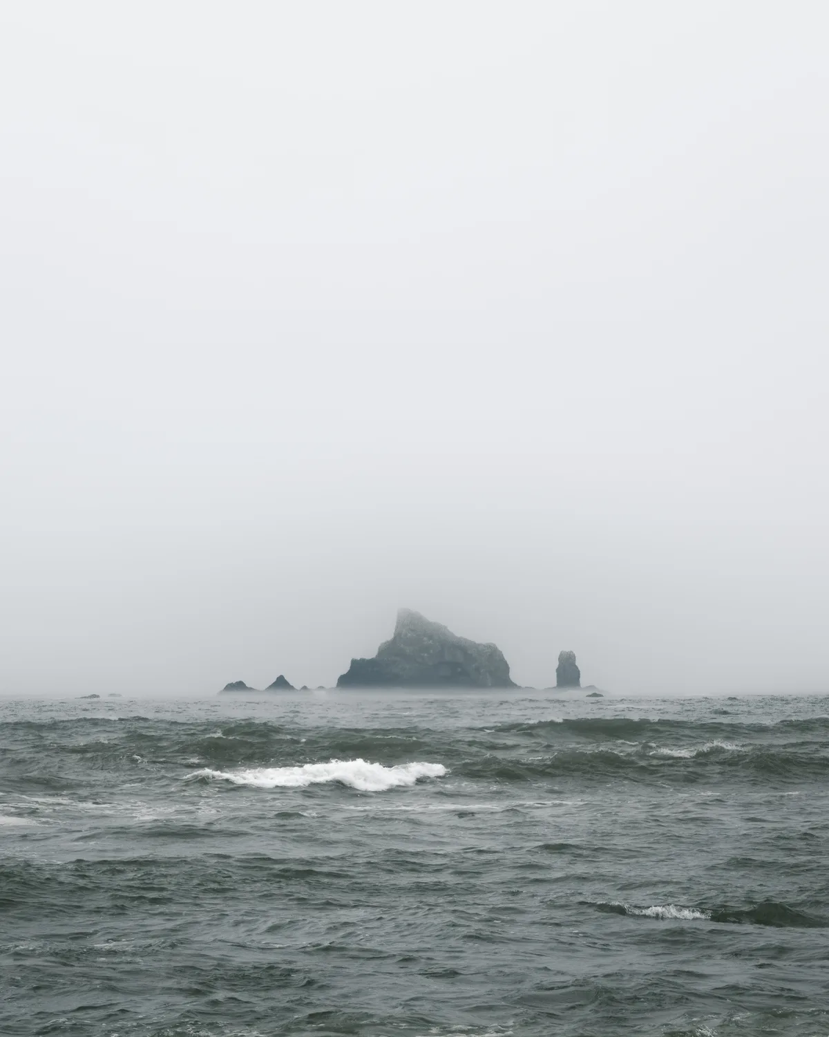 A lone sea stack rising from a grey, choppy ocean, half-dissolved in heavy coastal fog under a pale flat sky.