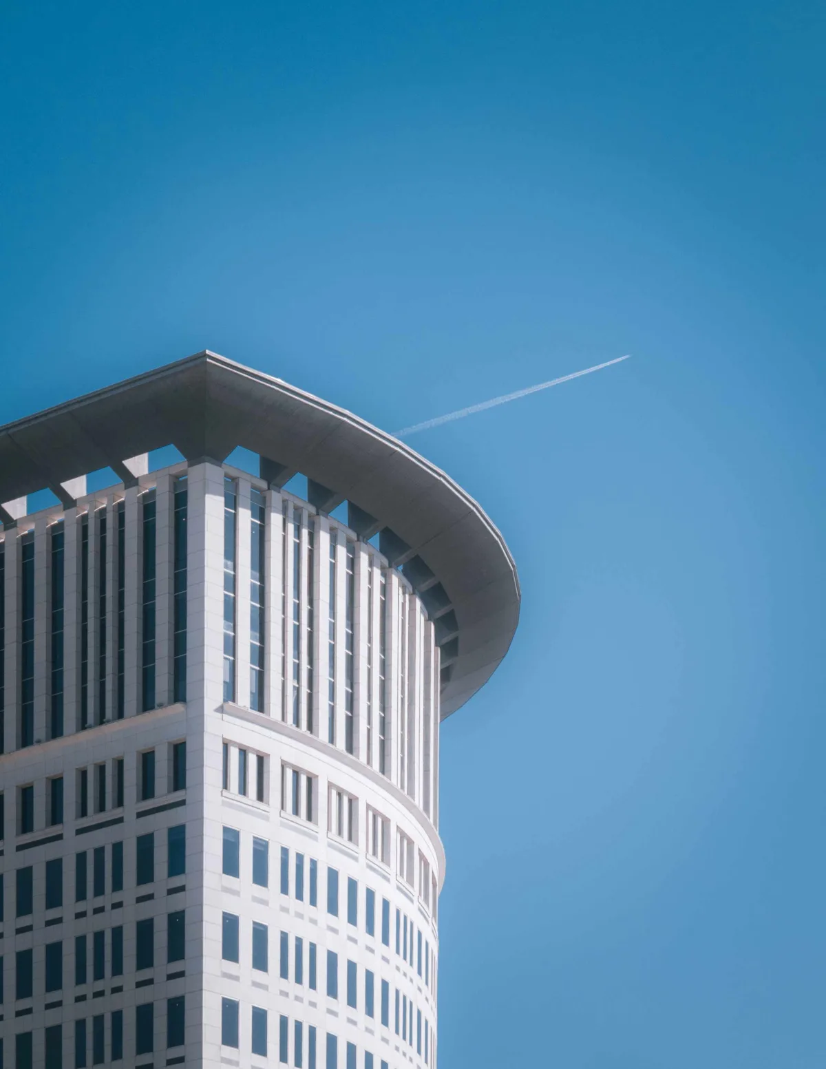The curved white upper floors of a modernist tower against a clear deep-blue sky, a single airplane contrail streaking past.