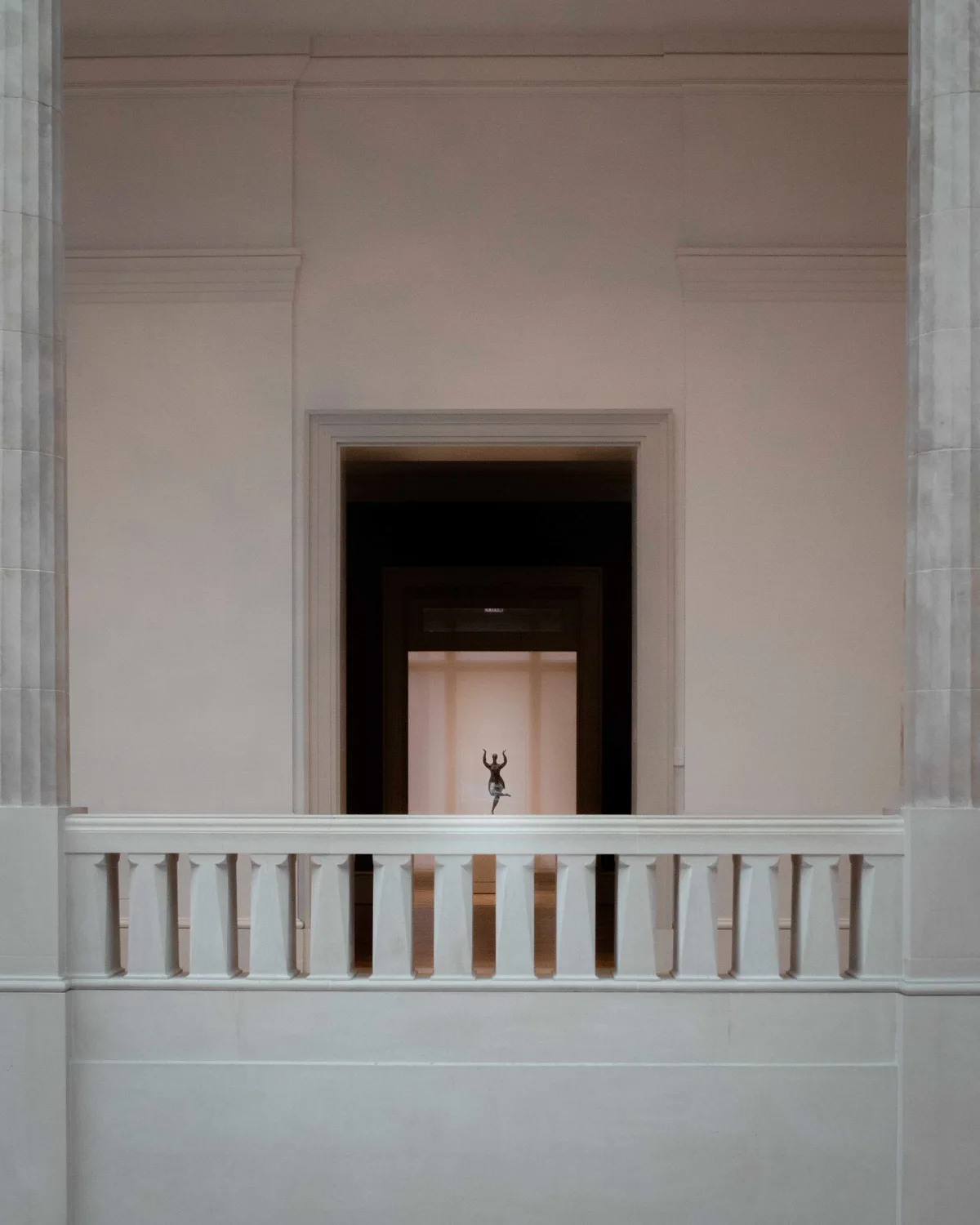 The pale marble interior of a grand hall seen over a balustrade, a small dancing figure framed in a distant lit doorway.