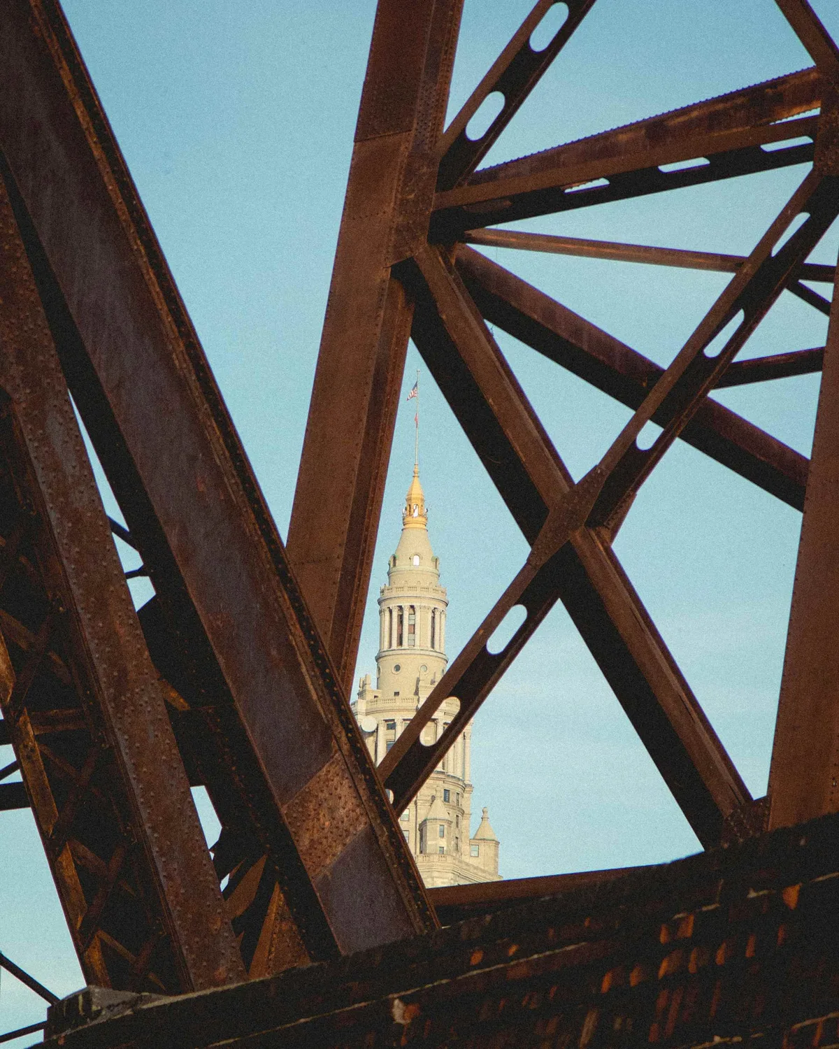 A gold-topped clock tower framed through the rusted steel girders of a bridge against a clear blue sky.