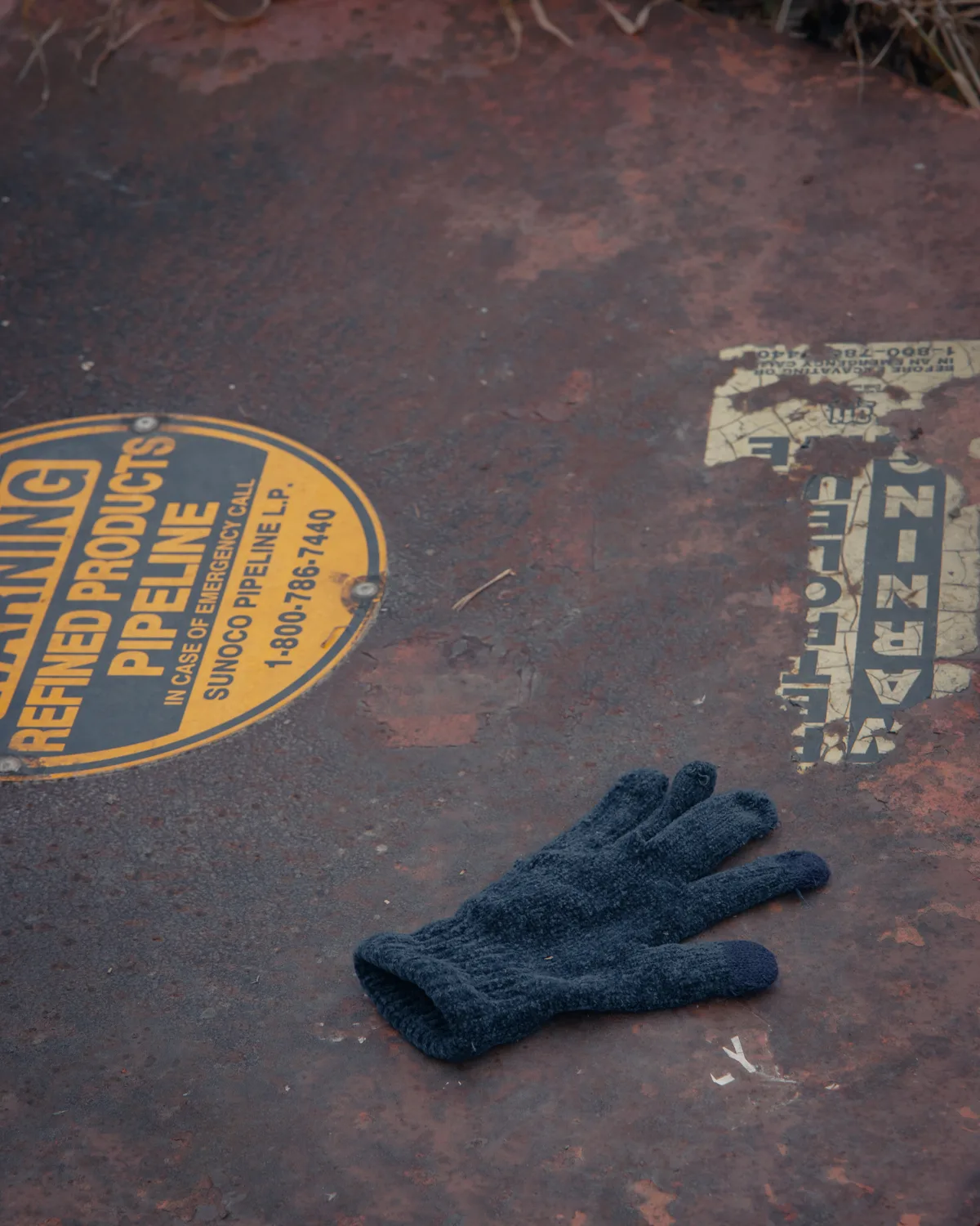 A child’s small navy knit glove lying on a rusted steel pipeline cover stamped with a yellow ‘Refined Products Pipeline’ warning.