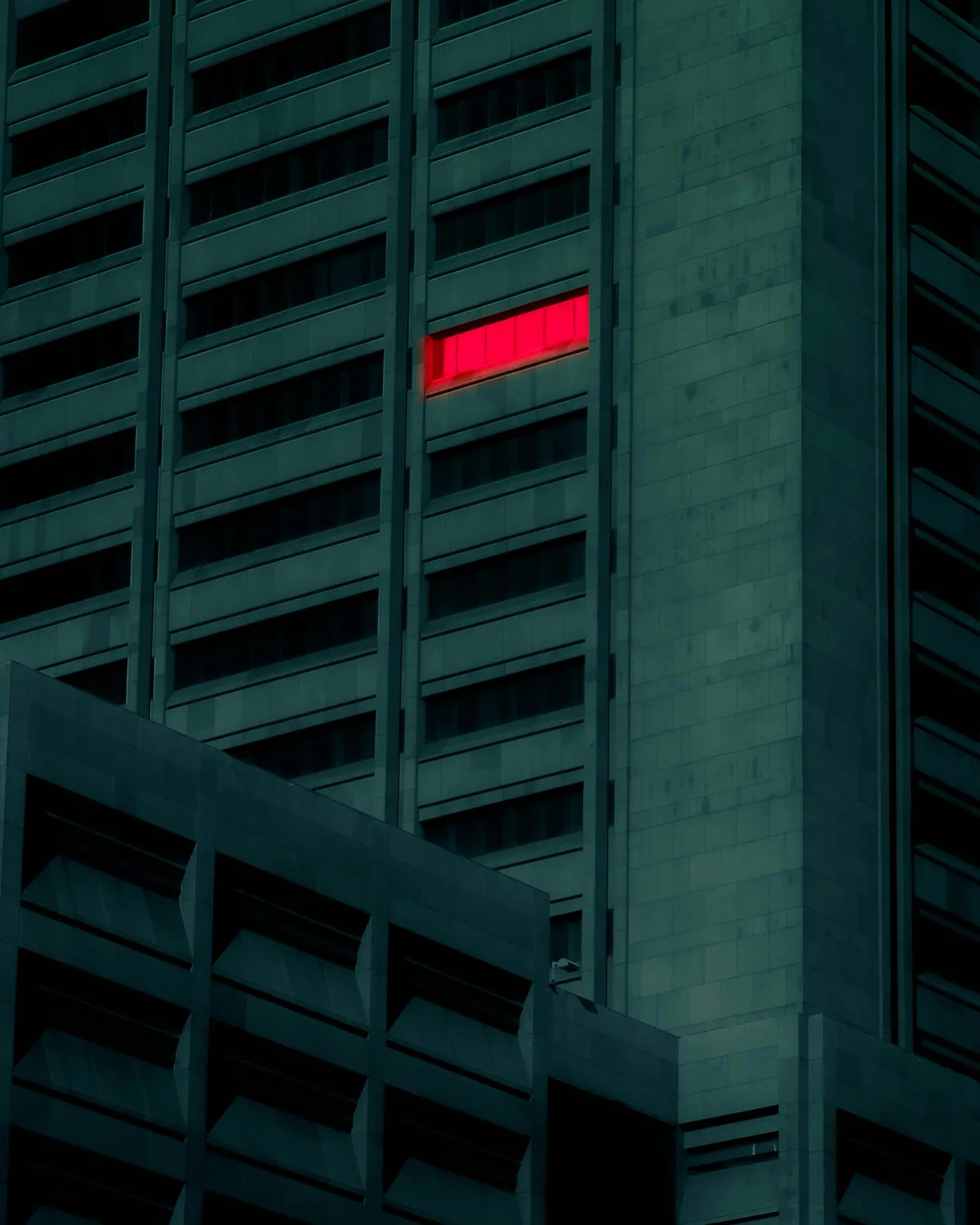 A dark teal office tower at dusk, every window unlit except one glowing red high on the facade.