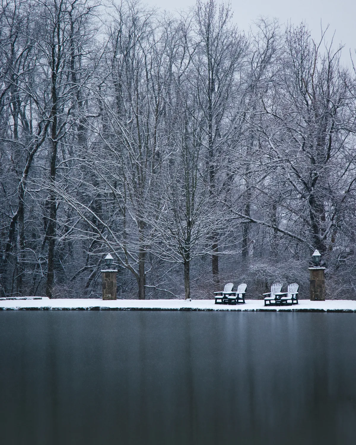 Two snow-dusted Adirondack chairs on the bank of a still, dark pond, snow-laden bare trees behind them in winter.