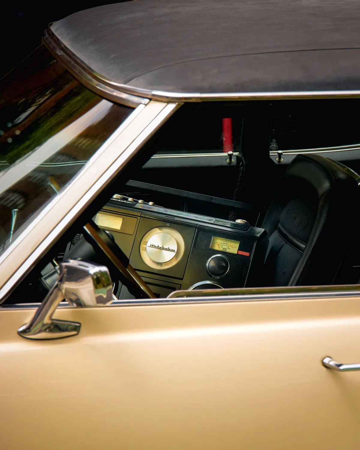 The interior of a vintage gold car seen through its side window, a chrome wing mirror in the foreground and an old dashboard radio within.