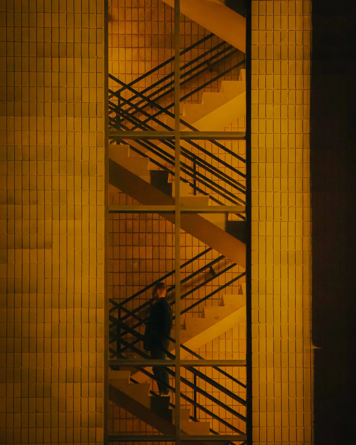 A lone figure climbing a zigzagging exterior staircase on a warmly lit tiled building, the flights doubling back above and below.